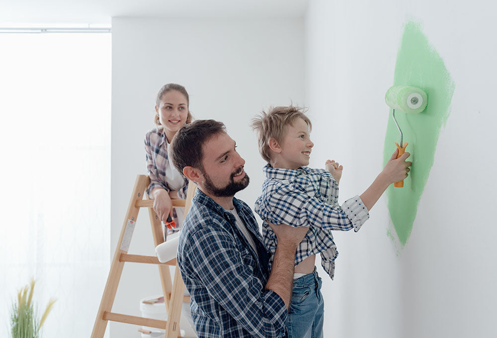 A father holding up a child to paint with a paint roller on the wall.